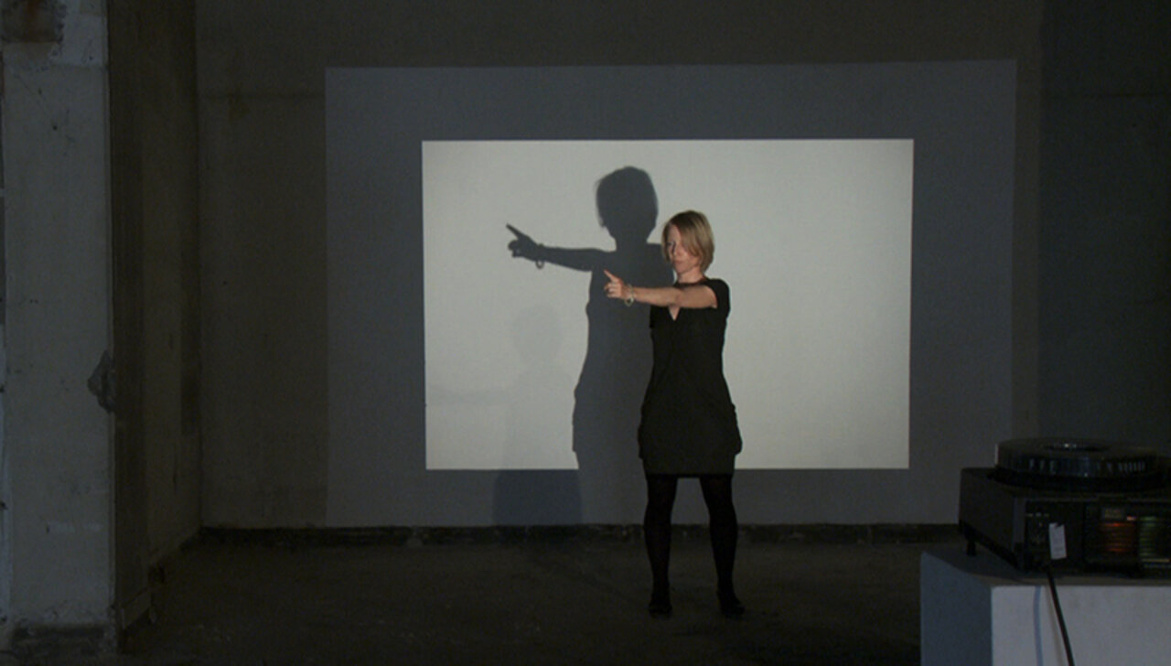 Carola Dertnig mit ausgestrecktem Arm und Zeigefinger vor einem Diaprojektor, der ihren Schatten an die Leinwand hinter ihr wirft